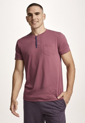 Lächelnder Mann, der ein kurzärmeliges mauve Henley-Shirt mit Brusttasche und gemusterten dunklen Hosen trägt, vor einem schlichten Hintergrund stehend.