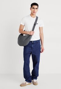 Sac à épaule à carreaux gris, en tissu doux, de forme courbée, porté par une personne en t-shirt blanc et jeans en denim bleu, avec des chaussures claires.