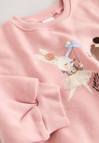 Sweat-shirt rose avec un graphisme de lapin accompagné d'une fleur, un ours et des accents en tulle. Texture douce et poignets côtelés pour plus de confort.
