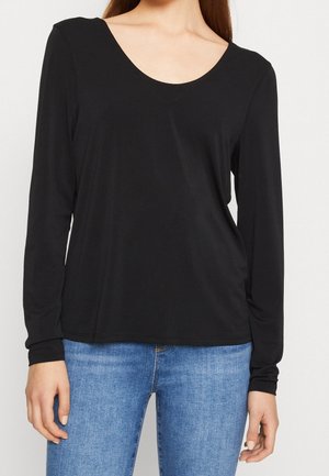 Kobieta ubrana w czarną bluzkę z długim rękawem i dekoltem w serek, w połączeniu z jasnoniebieskimi dżinsami, stojąca na jednolitym tle.