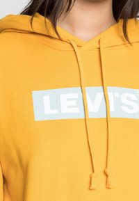 Osoba v hořčicově žluté mikině s kapucí, na které je bílé logo "LEVI'S" na světle modrém obdélníkovém pozadí, detail hrudníku a výstřihu.