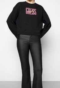 Sudadera negra corta con un prominente logo "GUESS" en rosa y amarillo, combinada con pantalones de campana negros brillantes. Tejido suave y texturizado.