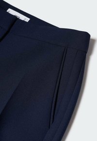 Námořnické modré kalhoty na míru s ostrými záhyby, vyrobené z hladkého materiálu. Mají plochý pas a skrytý zip.