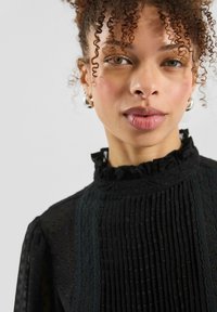 Zwarte blouse met een gerimpelde kraag, gestructureerde stof en verticale plooien. Voorzien van een stippenpatroon en subtiele kanten details.