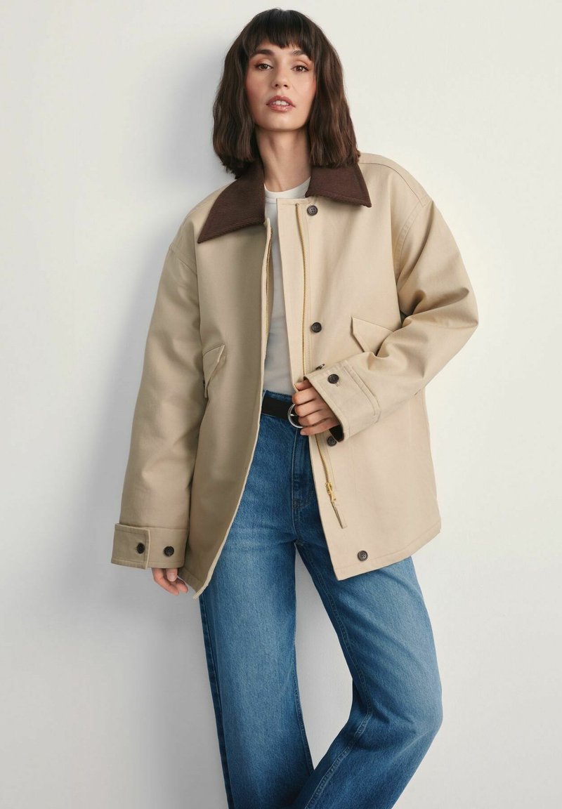 Kobieta w beżowej, oversize'owej kurtce z ciemnym kołnierzem, białej koszuli i niebieskich dżinsach, stojąca na tle jednolitego jasnego tła.