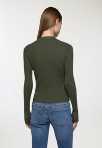 Oliwkowa, prążkowana bluzka z długim rękawem o dopasowanym kroju i okrągłym dekoltem, zestawiona z niebieskimi dżinsami z tylnymi kieszeniami.