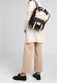 Personne portant un pantalon beige, des chaussures noires et un manteau clair, portant un sac à dos noir et beige dans le dos.