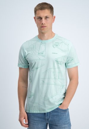 Jonge man met kort haar, gekleed in een lichtgroen T-shirt met patroon en blauwe spijkerbroek, staand tegen een effen achtergrond.