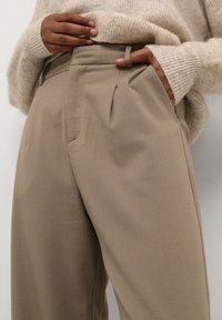 Persoon draagt beige broek met hoge taille, met één hand in de zak, en een beige gebreide trui waarvan de mouwen de polsen bedekken.