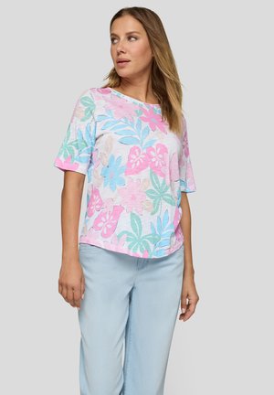 Frau trägt ein pastellfarbenes, kurzärmeliges florales Oberteil und hellblaue Hose, steht vor einem schlichten hellgrauen Hintergrund.