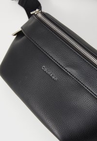 Zwarte leren heuptas met een gestructureerde afwerking, voorzien van een zilveren rits en een discrete Calvin Klein-logo dat op de voorkant is gedrukt.
