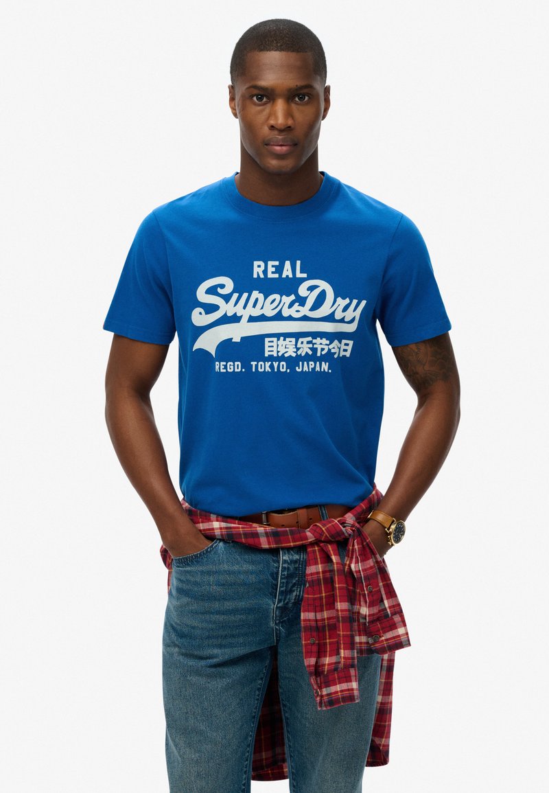 T-shirt en coton bleu avec un texte graphique blanc, associé à un jean bleu clair et une chemise à carreaux rouge nouée autour de la taille. Dispose d'un col rond.
