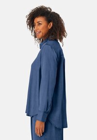 Donna con i capelli ricci che indossa una camicia azzurra a maniche lunghe con bottoni e pantaloni azzurri abbinati, di profilo con un leggero sorriso.