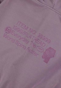 Fioletowa tkanina z wytłoczonymi tekstami i grafikami, w tym kodem przedmiotu, nazwą marki oraz stylizowanym wzorem spirali, wszystko w kolorze dopasowanym.