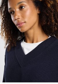 Navy knitted sweater with a deep V-neck design and ribbed texture, layered over a white shirt. Curly hair and natural skin tone visible.
