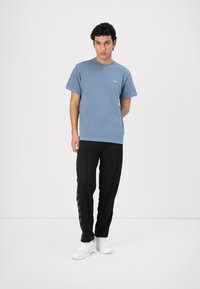 Blue cotton t-shirt with short sleeves and small text logo, paired with black athletic pants featuring a side stripe design and white sneakers.