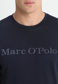 Meriini sininen puuvillat-paita, jossa on pyöreä kaula-aukko ja hopealla painettu brändinimi "Marc O’Polo" edessä rentoa fonttia käyttäen.