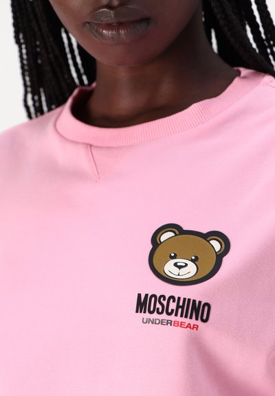Ružová mikina s grafickým motívom hnedého medveďa a textom "MOSCHINO UNDERBEAR" v čiernej a červenej farbe. Mäkká látka s rebrovaným výstrihom.