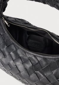 Musta nahkainen käsilaukku, jossa on kudottu muotoilu, avattu vetoketju paljastaa mustan kankaan sisätilan, jossa on pieni ommeltu etiketti.