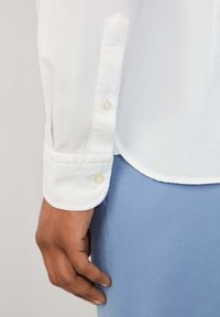 Hand met een gemiddelde huidskleur naast de manchet van een witte lange mouw shirt met knopen, boven een lichtblauwe broek.