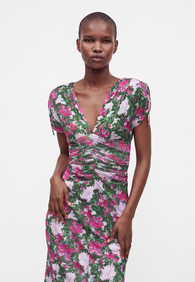 Femme aux cheveux courts portant une robe fleurie ajustée aux couleurs rose, blanche et verte, debout les mains sur les hanches devant un fond uni.