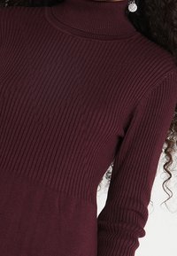 Femme portant un pull col roulé côtelé de couleur violet foncé avec des manches longues et des boucles d'oreilles rondes en argent.
