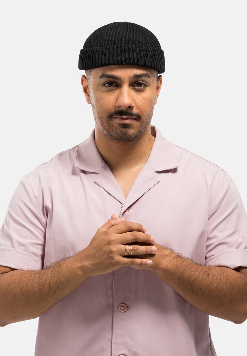 Gebreide zwarte beanie, lichtroze blouse met kraag en korte mouwen, drie gouden ringen op de handen, handen op borsthoogte samengevouwen, neutrale achtergrond.
