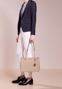 Béžová taška s ušima a zlatým řetízkem, hladká textura, detail loga, vyvážený obdélníkový tvar. Nosit ji osoba v tmavém saku a bílých džínách.