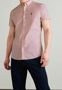 Korte mouwen, blouse met knopen in lichtroze stof met geborduurd zwart logo. Beschikt over een klassieke kraag en parelmoeren knopen.