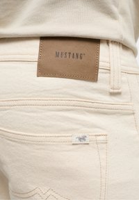 Jean en denim beige avec une étiquette en cuir marron portant l'inscription "MUSTANG". Détails de couture et petite étiquette avec un logo sur la poche arrière.