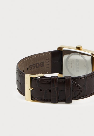 Bracelet de montre en cuir marron avec un motif crocodile, matériel doré, et un cadran carré avec un fond argenté. Gravure visible.