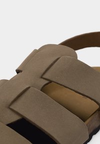 Sandale en daim marron avec de larges brides croisées et un bout ouvert, vue en gros plan sur un fond clair.