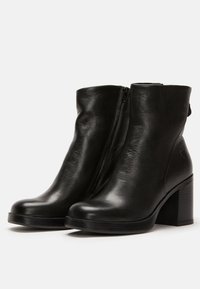 Botas de tobillo de cuero negro con una textura suave, que cuentan con un cierre lateral y un tacón grueso. Detalles notables incluyen un pequeño acento de logo.