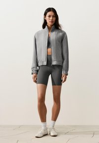 Femme debout portant une veste grise zippée, un soutien-gorge de sport gris foncé et un short de cycliste, des chaussettes blanches et des baskets blanches sur un sol clair.