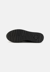 Semelle de chaussure en caoutchouc noir, présentant un motif de semelle multidirectionnel, une surface texturée et un logo de marque circulaire près du talon.