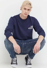 Felpa blu con logo "TOMMY JEAN WORLDWIDE", abbinata a jeans blu e sneakers alte nere, accessoriate con un cappello beige e un orecchino.