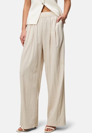 Femme portant un pantalon beige ample taille haute avec taille élastique, associé à des sandales ouvertes et un blazer sans manches, main dans la poche.