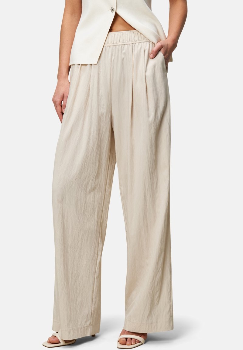 Femme portant un pantalon beige ample taille haute avec taille élastique, associé à des sandales ouvertes et un blazer sans manches, main dans la poche.