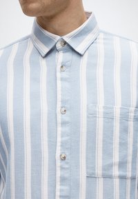 Γαλάζιο και λευκό πουκάμισο με ρίγες, κουμπώνει με κουμπιά, κατασκευασμένο από υφή υφάσματος, με κλασικό γιακά και τσέπη στο στήθος. Διαθέτει διάφανα πλαστικά κουμπιά.