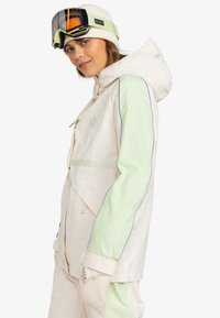 Letvægtsjakke i creme med mintgrønne ærmer, snoretræk til hætten og refleksdetaljer. Kombineret med beige vanter med et logo.