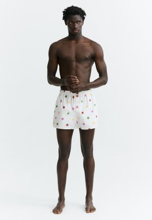 Giovane uomo in piedi a piedi nudi, indossa pantaloncini bianchi con pois colorati, senza maglietta, su sfondo bianco semplice.