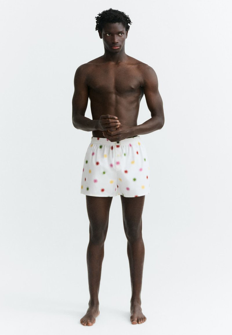 Jeune homme debout pieds nus portant un short blanc à pois colorés, torse nu, sur un fond blanc uni.