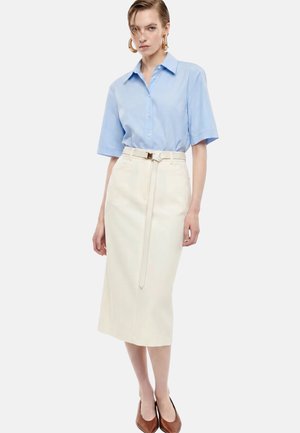 Donna che indossa una camicia azzurro chiaro a maniche corte con bottoni infilata in una gonna midi color crema con una cintura sottile e ballerine marroni con punta.