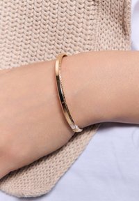 Złocona metalowa bransoletka w kształcie bangla, z gładką powierzchnią i teksturowanym wzorem, spoczywająca na dłoni na tle jasnej dzianiny.