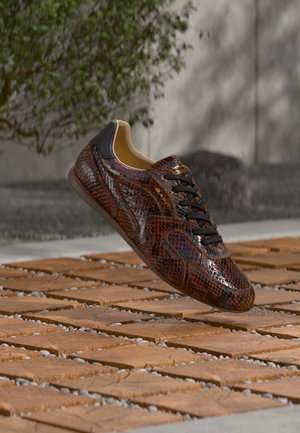 Barna és fekete kígyóbőr mintás sportcipő fekete fűzőkkel, mely fa lapok felett lebeg kültéren, háttérben elmosódott növények és szürke fal.