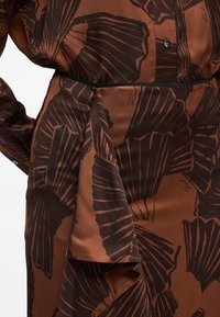 Brązowa tkanina w wzory z dużymi liśćmi w ciemniejszych odcieniach. Sukienka ma talię w stylu wrap z wiązanym detalem i czarnymi akcentami.