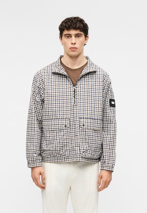 Ung mand iført en ternet lynlåstrøje med forlommer over en brun skjorte og hvide bukser, stående foran en ensfarvet baggrund.