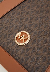 Smešani smeđa tkanina s uzorkom i reljefnim dizajnom, koja sadrži zlatni okrugli logo. Glatko svijetlosmeđe kožne akcentne detalje upotpunjuju dizajn proizvoda.
