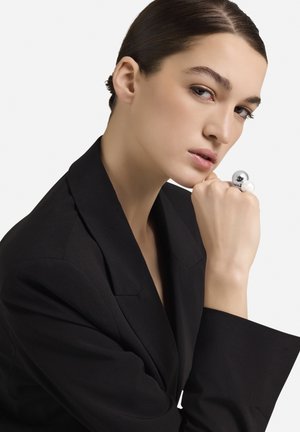 Juodas pritaikytas švarkas su plačiomis atlapomis, dėvimas modelio, palikusio smakrą ant rankos. Jos pirštą puošia blizgantis sidabrinis žiedas su perlais.