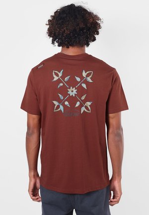 Homme aux cheveux bouclés portant un t-shirt marron avec un motif floral et le texte "Oxbow" au dos, debout devant un fond uni.